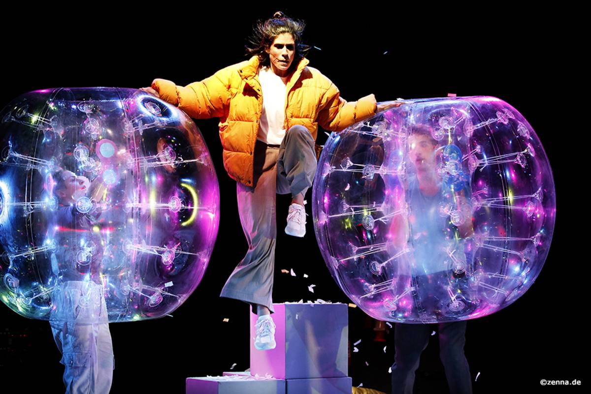 Zwei Tänzer:innen in großen Plastikkugeln rechts und links im Bild. In der Mitte eine Frau, die gerade in die Luft springt.