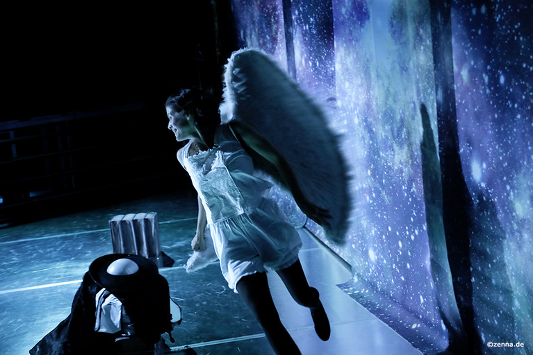 Eine Person mit weißem Overall und großen weißen Flügeln springt über die Bühne. Im Hintergrund ist ein Sternenhimmel zu sehen.