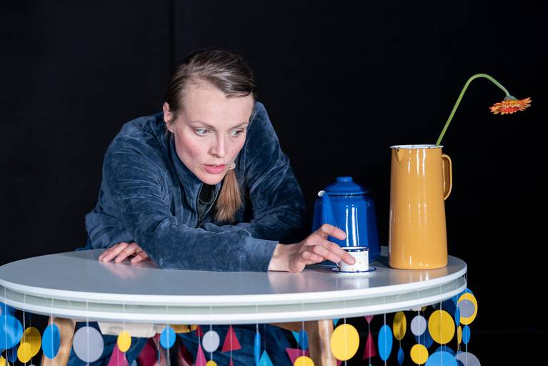 Inszenierungsfoto von "KLEIN". Schauspielerin Luise Audersch lehnt sich auf einen Tisch. Darauf stehen eine winzige Tasse, eine Teekanne und eine Blumenvase.