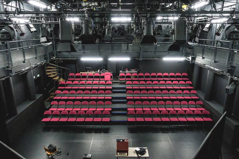 Der Publikumsbereich von der Bühne im Kleinen Saal aus. Eine Tribüne mit fünf Reihen mit je ungefähr zehn roten Stühlen links und rechts, dazwischen ein Durchgang. Die Bühne ist auf gleicher Höhe wie die erste Reihe.