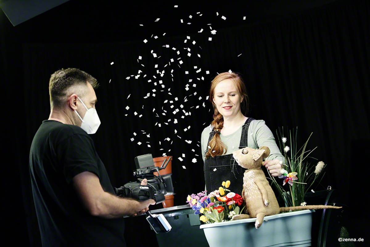 Schauspielerin Clara Fritsche hinten rechts im Bild mit der Mäuse-Figur. Vorne links im Bild ein Mann mit Kamera, der Clara filmt. Zusätzlich rieseln Blütenblätter herab.