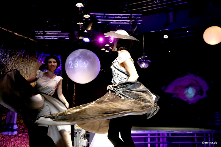 Zwei Menschen tanzen und tragen dabei weite Kleider und Hüte. Im Hintergrund sieht man einen lila Mond mit der Aufschrift: 23,9 Grad