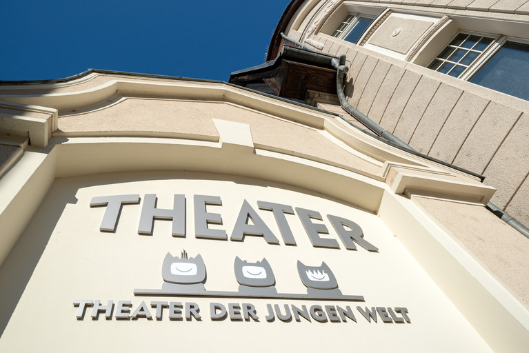 Die Schrift über dem Haupteingang des TDJWs in Nahaufnahme. Dort steht „Theater“, darunter die Köpfe von drei BOs. Darunter der Schriftzug „Theater der Jungen Welt“.