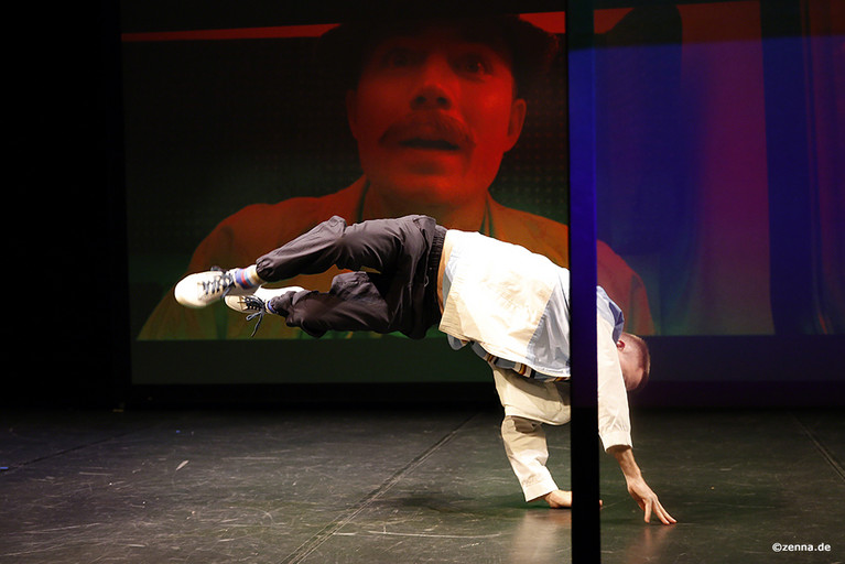 Schauspieler Benjamin Vinnen in einer Breakdance-Position. Hände sind auf dem Boden und Beine in der Luft. Im Hintergrund ist auf einer Leinwand ein dunkelrotes Bild von einem Mann mit großem Schnauzbart zu sehen.