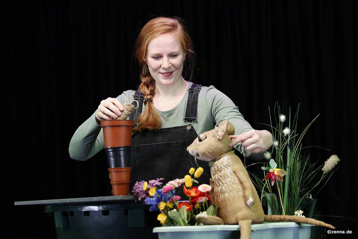 Puppenspielerin Clara Fritsche mit der Mäusefigur, die zwischen bunten Blumen sitzt.