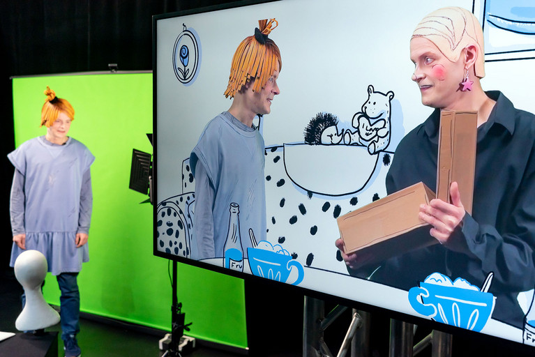 Schauspieler Sven Reese links im Bild vor einem Greenscreen verkleidet als Mädchen. Rechts im Bild ein großer Bildschirm mit Sven Reese als Mädchen und Sven Reese als Frau. Gemalte Umgebung ist ein Wohnzimmer.