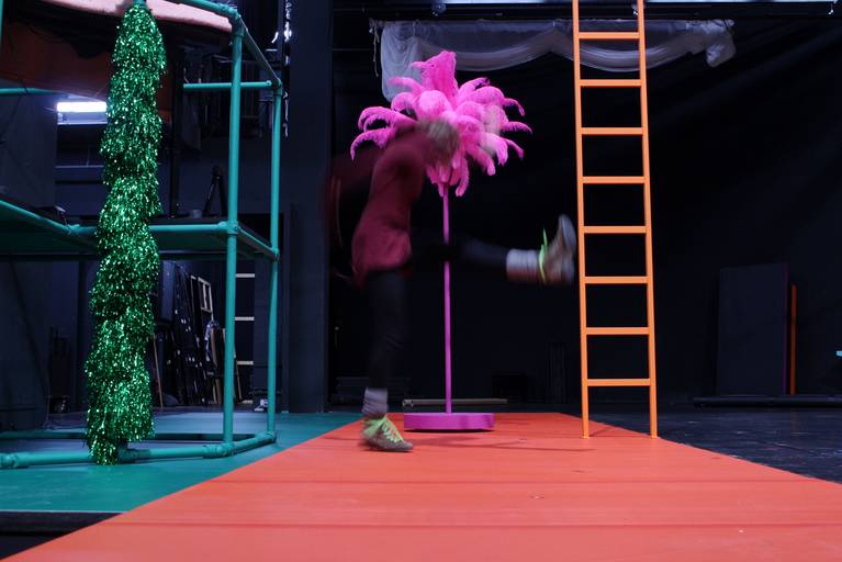 Bunte Bühnenkulisse mit grüner Girlande, pinker künstlicher Palme und orangener Leiter. In der Mitte des Bildes eine verschwommene Person, die ein Bein hebt.