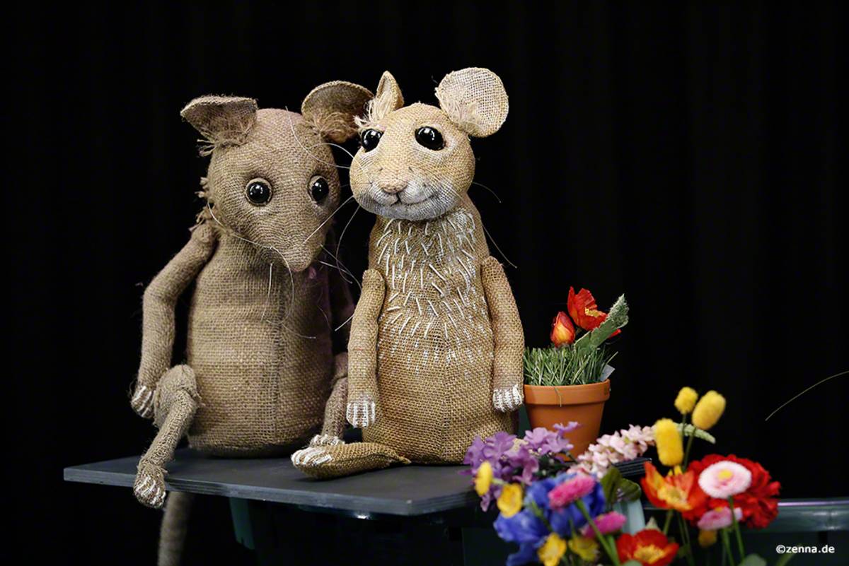 Zwei Mäuse-Figuren sitzen nebeneinander. Mit im Bild sind bunte Blumen.