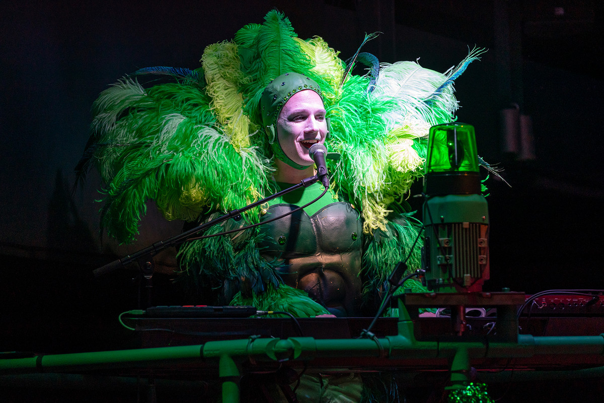 Schauspieler Benjamin Vinnen singend am Klavier in einem grünen Kostüm mit vielen Federn. 