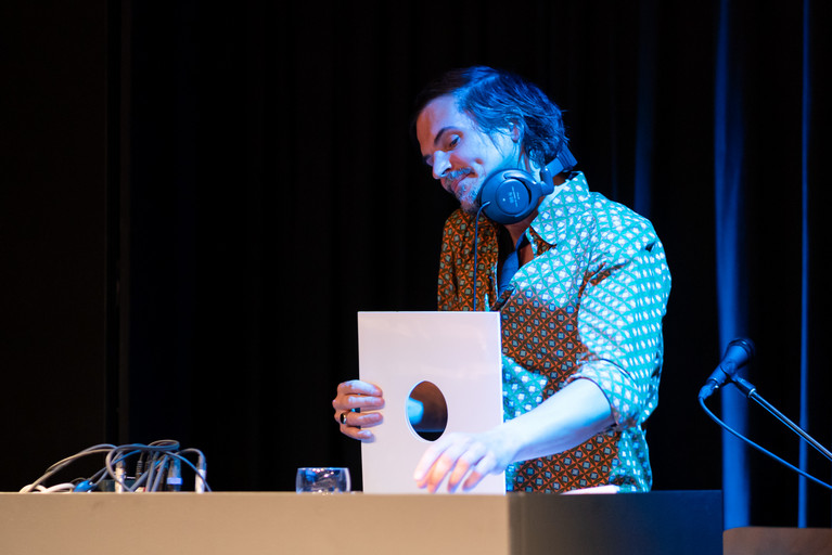 Eine Person steht hinter einem DJ-Pult und hält eine Vinyl-Platte in der Hand. Blaues Licht fällt auf die Szene.