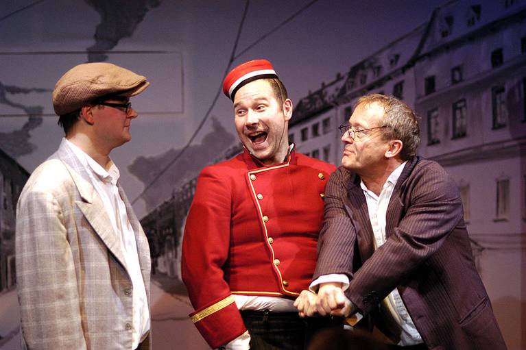 Inszenierungsfoto von "Emil und die Detektive". Zu sehen sind drei Männer. Der in der Mitte in einer roten Uniform.