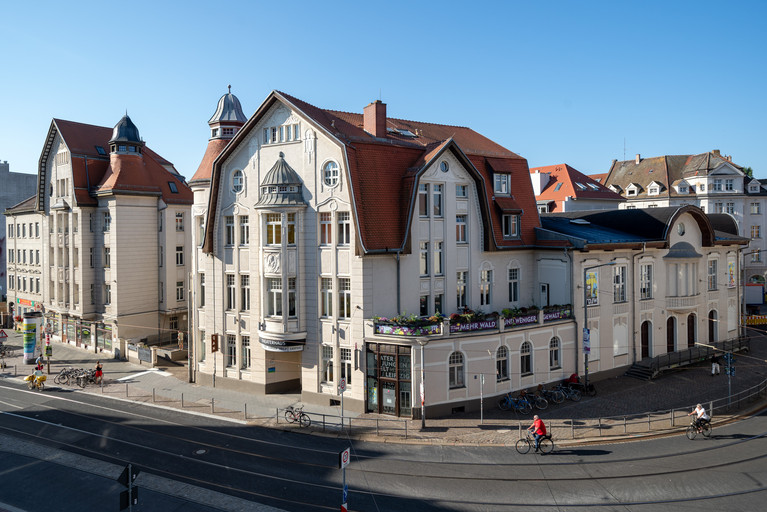 Das Gebäude des TDJWs von der Straßenecke Lindenauer Markt / Demmeringstraße. Es hat eine helle Farbe, ein rotes Dach und liegt an einer Straßenkurve. Abgebildet sind die Holztür zum Bühneneingang sowie die Glastür zum Ticketladen rechts daneben.