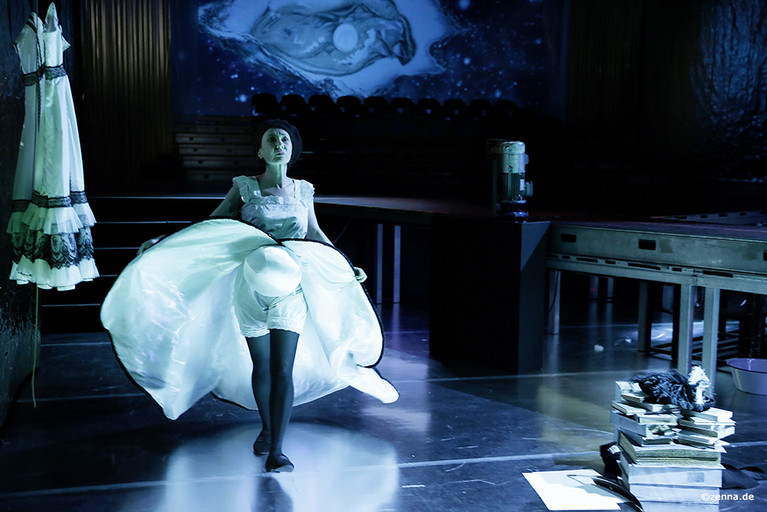 Eine Person mit weißem Kleid läuft neben einem Laufsteg entlang- Es fällt blaues Licht auf die Szene. In der rechten unteren Ecke liegt ein hoher Papierstapel.
