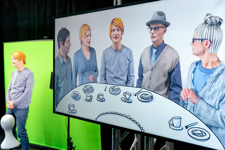 Schauspieler Sven Reese links im Bild vor einem Greenscreen. Rechts im Bild ein großer Bildschirm auf dem der Schauspieler zwischen mehreren Personen an einem Tisch sitzend zu sehen ist.